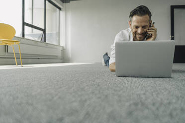 Lächelnder reifer Geschäftsmann auf dem Boden liegend in einem leeren Büro mit Laptop und Smartphone - KNSF06922