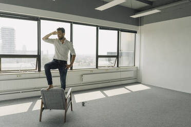 Mature businessman standing on armchair in empty office - KNSF06931