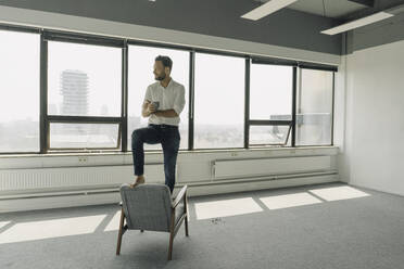 Mature businessman standing on armchair in empty office - KNSF06932