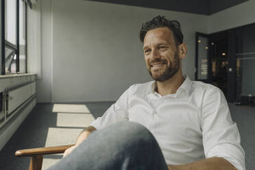 Smiling mature businessman sitting in armchair in empty office - KNSF06935