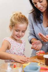 Mutter und Tochter beim Verzieren von Cupcakes mit Zuckerguss in der Küche - ISF23247
