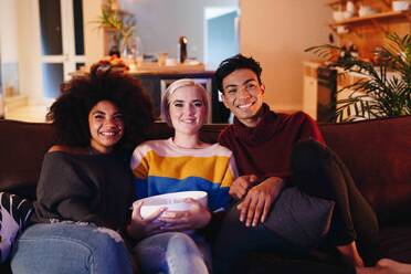 Junger Mann und junge Freundinnen sehen auf dem Wohnzimmersofa sitzend mit einer Schüssel Popcorn fern - ISF23327