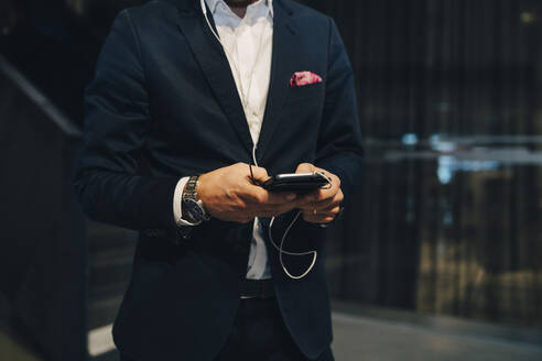 Mittelteil eines Geschäftsmannes, der ein Smartphone mit Kopfhörern im Büro benutzt - MASF15104