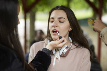 Teenage girl applying lipstick to female friend at park - MASF15113