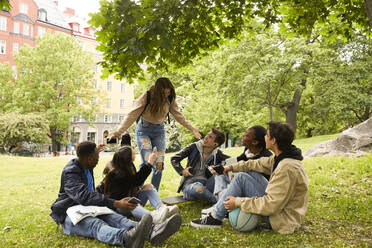 Fröhliches Mädchen im Teenageralter gestikuliert, während sie neben ihren Freunden steht, die im Park auf dem Boden sitzen - MASF15138