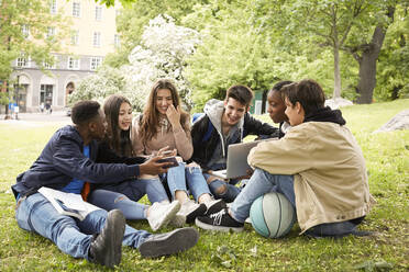 Smiling teenage boy showing smart phone to friends while sitting at park - MASF15139