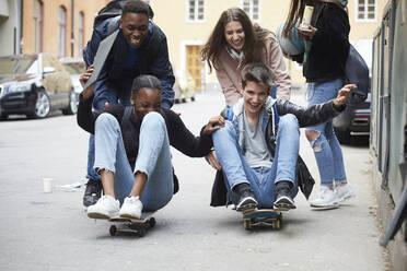 Cheerful friends pushing teenagers sitting on skateboard - MASF15160