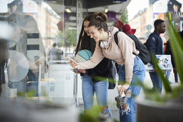 Lächelnde Freundinnen beim Fotografieren, während sie an einem Schaufenster in der Stadt stehen - MASF15174