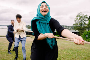 Lächelnde multiethnische männliche und weibliche Kollegen ziehen an einem Seil, während sie Tauziehen auf einer Wiese spielen - MASF15213