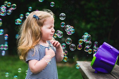 A little girl stands in front of a bubble machine. - CAVF69874