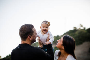 Vater hebt seine kleine Tochter hoch, während das Baby lacht und die Mutter zusieht - CAVF69900