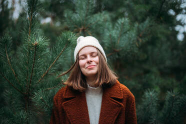Pretty woman in warm clothes standing against pine trees in forest - CAVF69954