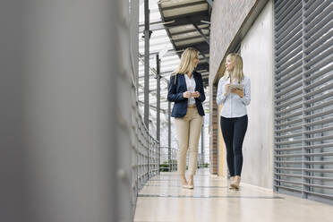 Two young businesswomen with tablet walking and talking in modern office building - JOSF03825