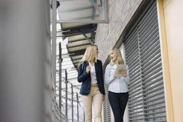 Two young businesswomen with tablet walking and talking in modern office building - JOSF03827