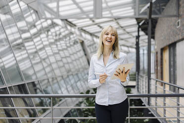 Lachende junge Geschäftsfrau in einem modernen Bürogebäude mit Tablet - JOSF03840