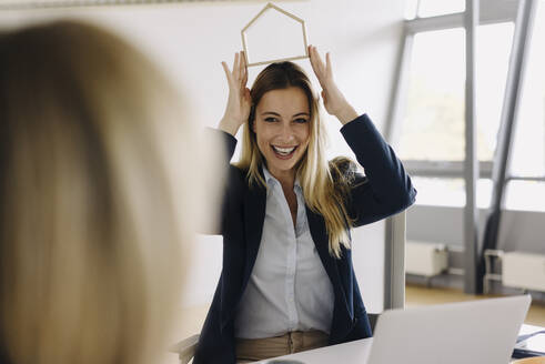 Glückliche junge Geschäftsfrauen zeigen Hausmodell am Schreibtisch im Büro - JOSF03852