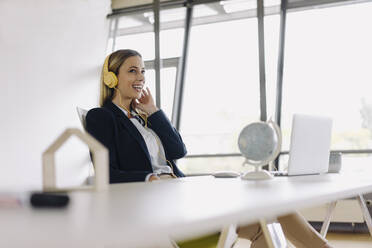 Happy young businesswoman with headphones and laptop at desk in office - JOSF03862