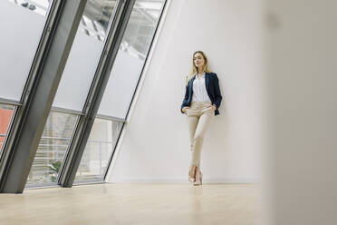 Young businesswoman leaning against a wall in a modern office building - JOSF03866