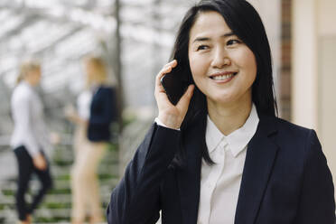 Portrait of a smiling businesswoman on the phone in office with colleagues in background - JOSF03882