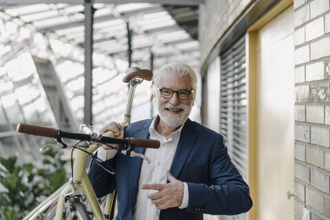 Porträt eines glücklichen älteren Geschäftsmannes mit einem Fahrrad in einem modernen Bürogebäude, lizenzfreies Stockfoto