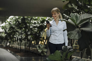 Smiling young businesswoman using cell phone in a modern office building surrounded by plants - JOSF04018