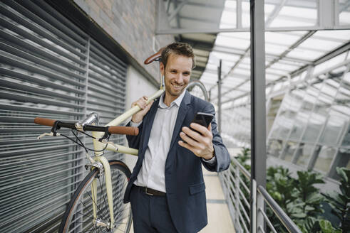 Lächelnder Geschäftsmann mit Mobiltelefon auf dem Fahrrad in einem modernen Bürogebäude - JOSF04032