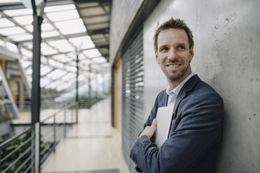 Smiling businessman leaning against a wall in modern office building - JOSF04038