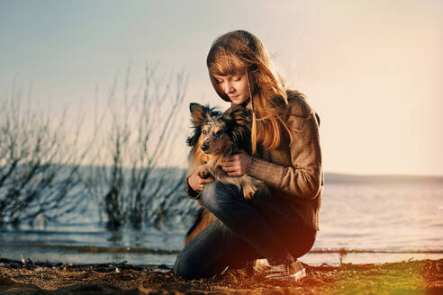 Teenager-Mädchen hält Hund, während sie bei Sonnenuntergang am Strand kniet - CAVF70049