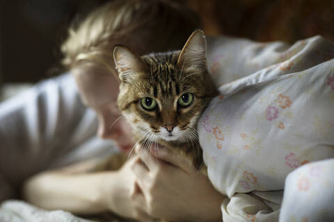 Frau umarmt Katze, während sie zu Hause auf dem Bett liegt, lizenzfreies Stockfoto