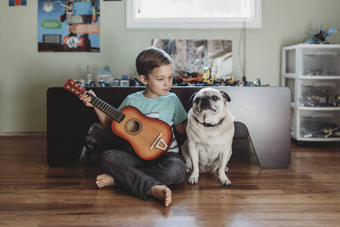 Barefoot boy holding guitar sitting next to pet Pug on hardwood floor - CAVF70083