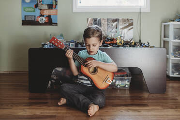 5 yr old boy sitting on hardwood floor playing guitar - CAVF70084