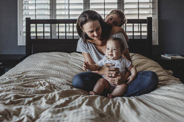 Glückliche Mutter auf dem Bett mit Baby auf dem Schoß und Sohn umarmt sie von hinten - CAVF70090