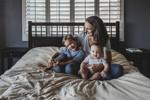 Lachende Mutter, die ihren kleinen Sohn und ihr Baby auf dem Bett in der Nähe des Fensters kuschelt - CAVF70091