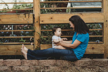 Lächelnde Mutter hält Baby auf ausgestreckten Beinen vor einem Zaun - CAVF70098