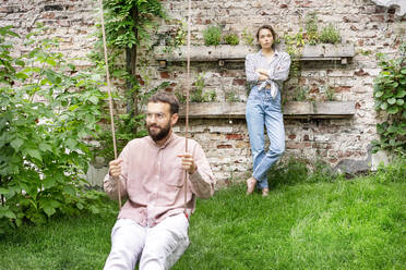 Paar im Garten, Mann sitzt auf einer Schaukel, Frau lehnt sich an die Wand und schaut verärgert - PESF01736
