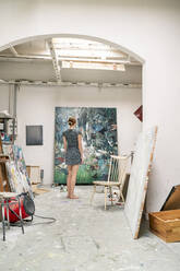 Junge Künstlerin steht in ihrem Atelier und betrachtet ein Gemälde, Rückansicht - PESF01741