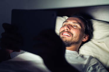 Junger gutaussehender und müder Mann mit Bart lächelt nachts auf ein Tablet - CAVF70120