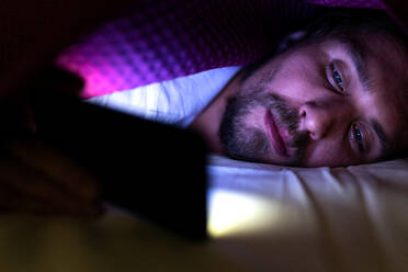 Bärtiger junger Mann liegt im Bett unter seiner Decke und schaut auf sein Handy - CAVF70123