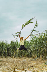 Ein Bauer mit Hut springt in einem grünen Feld und hält Maisstängel hoch. - CAVF70141
