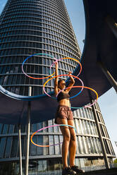 Active woman performing Hula Hoop dance with five rings in urban area - CAVF70194