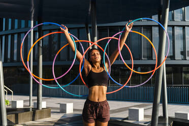 Generation Z young woman holding five Hula Hoop in downtown at sunset - CAVF70195