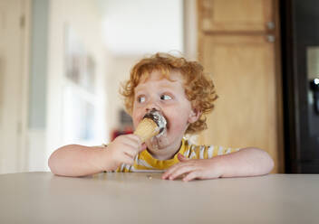 Messy toddler boy eats ice cream while sitting at counter in kitchen - CAVF70252