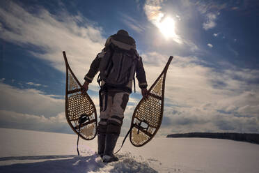 Rückansicht eines Mannes mit Rucksack, der Schneeschuhe trägt, während er über ein verschneites Feld läuft - CAVF70276