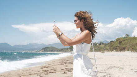 Side view of woman photographing through smart phone while standing at shore during summer - CAVF70283