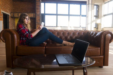 Side view of young woman using tablet computer while sitting on leather sofa at home - CAVF70293