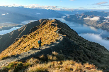 Man walking along ridge line toward peak of mountain overlooking lake. - CAVF70342