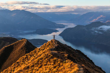 Man standing at peak of mountain overlooking blue lake during sunset. - CAVF70344