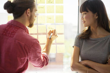 Businesswoman looking at colleague writing on adhesive note in creative office - CAVF70428