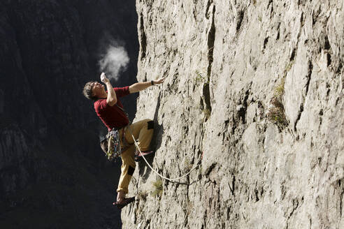 Männlicher Kletterer, der eine Felswand erklimmt, nach oben schaut und Kreide auf die Hände pustet - HOXF04493