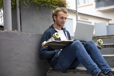 Young man sitting on steps, using laptop on longboard - MOEF02707